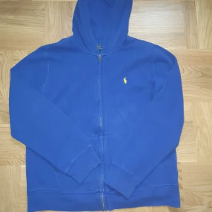 Blå hoodie från Polo Ralph Lauren - Blå hoodie från Polo Ralph Lauren med dragkedja och huva. Klassisk gul broderad logga på bröstet. Mjuk och skön, perfekt för chill dagar. Långärmad modell med ribbade muddar och fickor framtill.