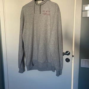 Grå hoodie med tryck Je m'en amour - Grå hoodie med huva och snörning, tryck i rosa och svart med texten 'Je m'en amour' både fram och bak. Mjuk och skön, perfekt för chill dagar. Långärmad och i klassisk passform. Materialet känns som bomullsmix. Den är i storlek 170 men är stor i modellen 