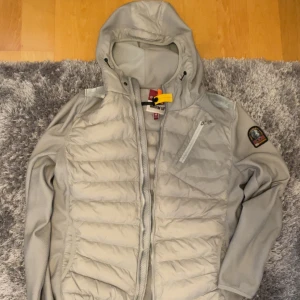 Ljusgrå pufferjacka från Parajumpers - Snygg ljusgrå pufferjacka från Parajumpers med huva, dragkedja och coola detaljer som bröstficka och patch på ärmen. Jackan har quiltad framsida och är tillverkad i syntetmaterial. Perfekt för dig som vill ha en stilren och sportig look.