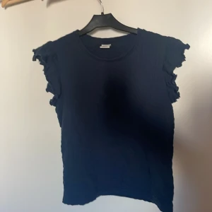 Mörkblå volangtopp från Lindex - Supersöt mörkblå topp från Lindex med volangdetaljer på ärmarna. T-shirten har rund halsringning och är tillverkad i ett mjukt, lätt strukturerat bomullstyg. Perfekt för dig som gillar en enkel men ändå lekfull stil.