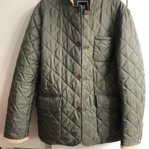 Quiltad grön jacka Hansen & Jacob - Quiltad grön jacka från Hansen & Jacob med snygg manchesterkrage i beige. Jackan har stora fickor framtill, knappar med logga och ribbade muddar. Perfekt för dig som gillar klassisk stil med modern twist. Ny /Oanvänd