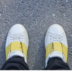 Valentino Open sneakers vit/gul - Säljer ett par Valentino Open sneakers i vitt skinn med bred gul läderdetalj över vristen. Klassisk rund tå, vita snören och platt sula med små nitar på hälen. Perfekta för dig som vill sticka ut med en färgklick i din outfit.