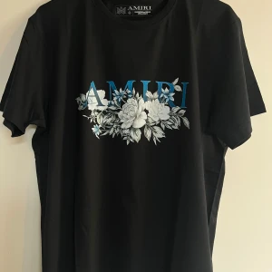 Svart Amiri t-shirt med blommor - Svart t-shirt från Amiri med stor blå logga och detaljerat blommönster i vitt och grått på bröstet. Klassisk passform och rund halsringning. Perfekt för dig som vill sticka ut med en stilren men ändå unik look.