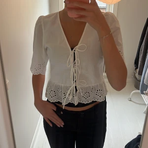 Vit blus med snörning och broderi - Superfin vit blus med v-ringning och snörning framtill. Blusen har korta ärmar och broderade blommor längs ärmslut och nederkant. Modellen är lite mer croppad. Aldrig använda i storlek XS från Gina.
