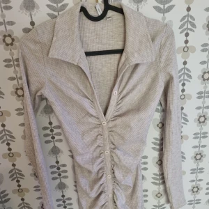 Beige ribbad klänning  - Trendig beige klänning från nelly med ribbat tyg och snygg rynkdetalj längs knappraden. Djup v-ringning, stora kragar och långa ärmar ger en cool och modern look. Perfekt för dig som gillar stilrena men unika plagg.