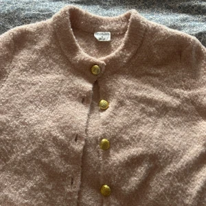 Beige kofta från Stories med guldknappar - Superfin beige kofta från Stories i storlek S. Koftan är gjord i ett mjukt, stickat material och har fem snygga guldknappar framtill. Rund halsringning och två fickor framtill ger en chill och trendig vibe. Perfekt att slänga på sig när det är lite kyligt ute.