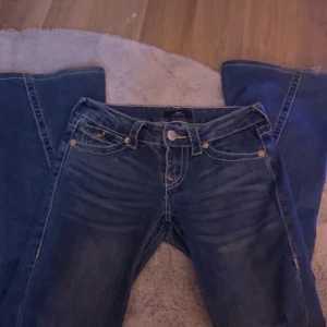 True Religion Joey Low Rise Flare Jeans - Säljer ett par blå True Religion Joey jeans med låg midja och flare-ben. Klassiska kontrastsömmar, snygga detaljer på bakfickorna och guldiga knappar. Jeansen är i bomull och har en cool vintagekänsla. Perfekta för dig som gillar statement-jeans!