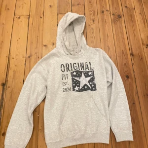 Grå hoodie från Gildan med tryck - Säljer en ljusgrå hoodie från Gildan med trycket 'ORIGINAL FIT EST. 2024' och en stor stjärna med mindre stjärnor på bröstet. Tröjan har huva med snörning och en klassisk känguruficka framtill. Perfekt för chill dagar.
