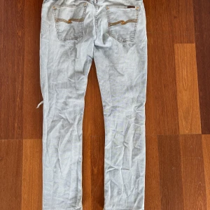 Ljusblå/grå Nudie Jeans 32/32 - Säljer ett par ljusblågråa jeans från Nudie Jeans med klassiska orange sömmar och broderade bakfickor. Modellen har rak/slim passform, samt slitning och hål på ena knät för en cool, avslappnad look. 