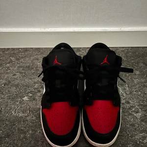 Nike Air Jordan 1 Low svart/röd/vit - Säljer ett par Nike Air Jordan 1 Low i klassiska färger: svart, röd och vit. Skorna har svart snörning, vit sula och den ikoniska Air Jordan-loggan broderad på hälen. Ovandelen är i skinn med perforerad tåbox och röda detaljer. Perfekta för dig som gillar streetstyle.