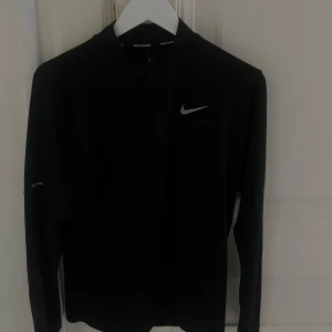 Svart Nike långärmad träningströja - Svart långärmad träningströja från Nike med halv dragkedja framtill och klassisk Nike-logga på bröstet. Perfekt för löpning eller gym, tillverkad i ett mjukt och stretchigt syntetmaterial som andas. Snygg och sportig design med tight passform.