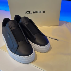 Svarta sneakers från Axel Arigato - Stilrena svarta sneakers från Axel Arigato i skinn med vit platt sula. Minimalistisk design utan snörning, elastiskt band över vristen och diskret logga i guld på sidan. Perfekta för dig som gillar clean och modern look. Allt i bilden ingår. Nypris 2 900kr. Mitt pris 1200kr. HELT nya, oanvända.  Skriv om du vill ha mer information! 👌🏼