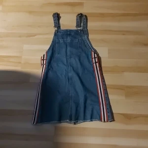 Jeansklänning med ränder från H&M Divided - Säljer en blå jeansklänning från H&M Divided i storlek 38. Klänningen har hängslen, en ficka framtill och coola röda och vita ränder längs sidorna. Tillverkad i bomullsmix med lite stretch för skön passform. Perfekt för en avslappnad och trendig look!