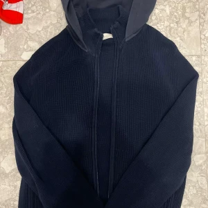 Mörkblå stickad cardigan från Sayless - Mörkblå stickad hoodie från Sayles i storlek XS. Tröjan har huva, dragkedja framtill och är ribbstickad för en snygg struktur. Perfekt för dig som gillar en enkel men stilren look med lite sportig känsla.