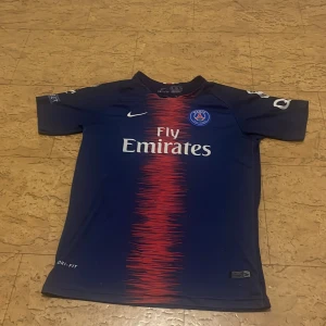 PSG Mbappé matchtröja Nike Dri-Fit - Säljer en officiell Paris Saint-Germain fotbollströja med Mbappé och nummer 7 på ryggen. Tröjan är marinblå med en röd randig effekt i mitten, vita detaljer och PSG-logga på bröstet. Tillverkad i lätt Dri-Fit material som andas, perfekt för träning eller match.