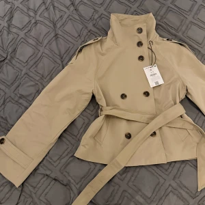Beige trenchcoat med bälte, S - Stilren beige trenchcoat med dubbelknäppning, höga axelklaffar och bälte i midjan. Jackan har långa ärmar med knappar vid ärmslut och klassisk krage. Perfekt för dig som gillar en clean och tidlös look.  