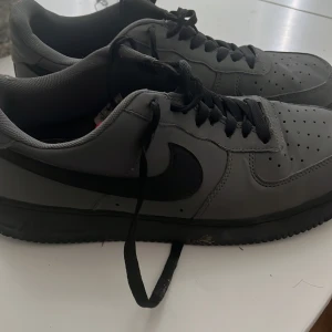 Gråa Nike Air Force 1 sneakers - Snygga grå Nike Air Force 1 sneakers med svart swoosh och svarta snören. Skorna har klassisk rund tå, perforerade detaljer på tån och platt sula. Perfekta för dig som gillar stilrena och sportiga sneakers.