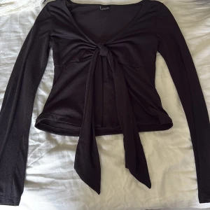 Svart långärmad knytblus Gina Tricot - Svart långärmad topp från Gina Tricot i storlek XS. Modellen har v-ringning och en snygg knytning framtill som ger en cool och trendig look. Tunt och mjukt material som sitter slim på kroppen. Perfekt att styla med jeans eller kjol.