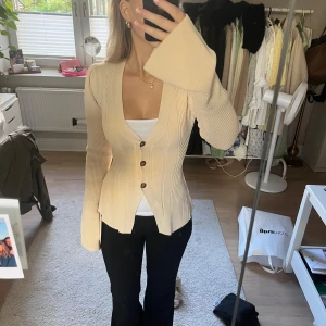 Helt ny med prislapp. Beige ribbad kofta med vida ärmar - Säljer en beige ribbad kofta med markerad midja och vida, långa ärmar. Koftan har djup v-ringning och knäpps med tre bruna knappar framtill. Materialet är stickat och stretchigt, perfekt för lager-på-lager och snygg till jeans.