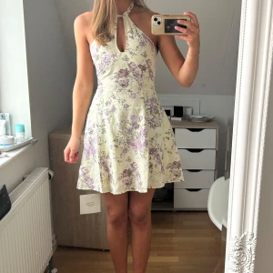Blommig halterneckklänning i ljusa toner - Supersöt kort klänning med halterneck och nyckelhålsdetalj framtill. Klänningen är ljus med lila och grått blommönster, har en utsvängd kjol och är ärmlös. Perfekt för varma dagar och somriga tillfällen. Säljer då den är lite liten på mig. Går inte att köpa längre på hemsidan 