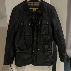 Svart quiltad jacka från Belstaff - Snygg svart quiltad jacka från Belstaff med bälte i midjan och flera fickor framtill. Jackan har dragkedja, tryckknappar och klassisk krage. Insidan har rutigt foder och jackan är perfekt för höst och vinter. Stilsäker och tidlös design.