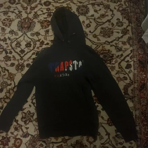 Svart Trapstar hoodie med tryck - Säljer en svart hoodie från Trapstar med färgglatt tryck i blått, rött och vitt på bröstet där det står 'Trapstar It's A Secret'. Hoodien har huva med snörning och är långärmad. Perfekt för dig som gillar streetwear och vill sticka ut. Priset går att diskuteras vid snabb affär