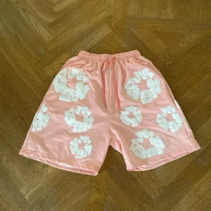 Denim tears - Snygga rosa shorts från Denimtears i storlek S med elastisk midja och snörning. De har ett unikt vitt mönster över hela tyget och en bakficka. Materialet känns mjukt och lätt, perfekt för varma dagar. Passformen är loose och de har en chill vibe.