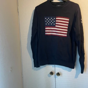 Mörkblå GANT tröja med USA-flagga - Säljer en mörkblå långärmad tröja från GANT med en stor amerikansk flagga broderad på bröstet. Tröjan har ribbade muddar vid ärmslut och nederkant samt diskret tryck på ena ärmen. Perfekt för dig som gillar klassisk college-stil.
