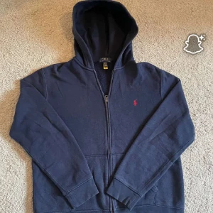 👕 Polo Ralph Lauren Hoodie - 👕 Polo Ralph Lauren Hoodie Stilren marinblå hoodie i klassisk design ✨. Dragkedja framtill 🔗, huva med dragsko 🧢 och praktiska kängurufickor 👐.  ✔️ Mjukt och bekvämt bomullsmaterial – perfekt för vardag & fritid ✔️ Ikonisk röd Polo-logga 🐎 broderad på bröstet ✔️ Ribbade muddar vid ärmslut & nederkant för bra passform 💯  Passar lika bra till jeans 👖 som joggers 👟. Ett tidlöst basplagg som aldrig går ur stil – både snyggt och bekvämt! 🙌