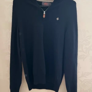 Mörkblå merinoullströja från Morris - Säljer en stilren mörkblå tröja från Morris i 100% merinoull. Tröjan har en liten broderad logga på bröstet, halv dragkedja med läderdetalj och lång ärm. Perfekt för dig som gillar klassisk och enkel stil.