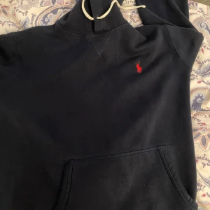 Mörkblå hoodie från Polo Ralph Lauren - Svart hoodie från Polo Ralph Lauren med klassisk röd broderad logga på bröstet. Tröjan har huva med vita snören och en stor magficka. Perfekt för dig som gillar stilren streetwear med premiumkänsla.