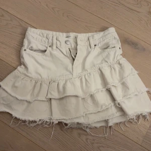 Vit volangkjol i jeans från Gina  - Supersöt vit kortkjol i jeans från Gina med flera volanger och råa kanter. Kjolen har klassiska jeansdetaljer som fickor och bälteshällor, perfekt för en trendig och avslappnad look.