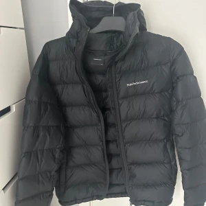 Svart dunjacka Peak Performance - Säljer en svart dunjacka från Peak Performance med huva och dragkedja. Jackan är quiltad och har en stilren look med logga på bröstet. Perfekt för kalla dagar och enkel att matcha med allt. Snygg och funktionell för vintern!