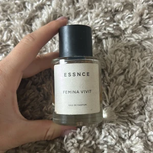 ESSNCE Femina Vivit Eau de Parfum - Minimalistisk parfymflaska från ESSNCE med doften Femina Vivit. Flaskan är rund i genomskinligt glas med svart cylinderformad kork och vit etikett med svart text. Perfekt för dig som gillar stilren design och unika dofter.
