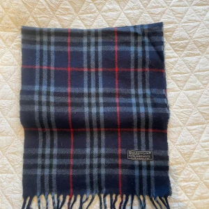 Rutig halsduk från Burberry - Säljer en klassisk rutig halsduk från Burberry i mörkblått med röda och grå ränder. Tillverkad av 100% lammull, vilket gör den både mjuk och varm. Perfekt accessoar för kyliga dagar. Märkt med 'Made in England'. Säljer då jag inte använder den så mycket. Skicket är 9/10. 