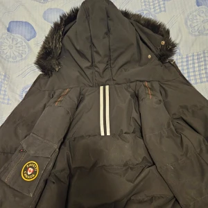 Svart pufferjacka från Zavetti Canada - Säljer en svart pufferjacka från Zavetti Canada med pälsfodrad huva och broderad logga på ärmen. Jackan har dragkedja och knappar framtill samt quiltad insida. Perfekt för kalla dagar! Storlek L i barn/160