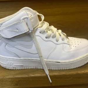 Vita Nike Air Force sneakers - Snygga vita Nike Air Force sneakers med hög modell och klassisk snörning. Skorna har en justerbar rem vid ankeln för extra stöd och stil. Perfekta för en sportig look.