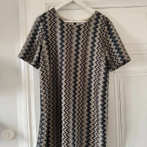 Mönstrad klänning från Drylake - Snygg klänning med zigzag-mönster i svart, beige och grå toner. Den har korta ärmar och en dragkedja i ryggen. Perfekt för en stilren look. Missoni-inspirerad.
