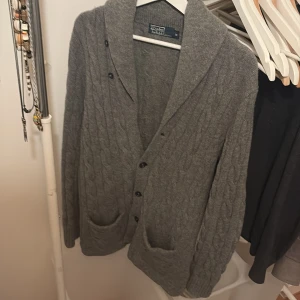 Grå cardigan från Polo Ralph Lauren - Säljer en stilren grå cardigan från Polo Ralph Lauren. Den är kabelstickad med knappar framtill och har två praktiska fickor. Perfekt för kyliga dagar med sitt mjuka material och bekväma passform. Storlek: M passar L. En knapp saknas.