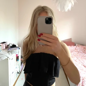 Svart axelbandslös topp - Snygg svart axelbandslös topp med volangdetaljer. Perfekt för en kväll ute eller en speciell tillställning. Toppen har en elastisk baksida för bättre passform.