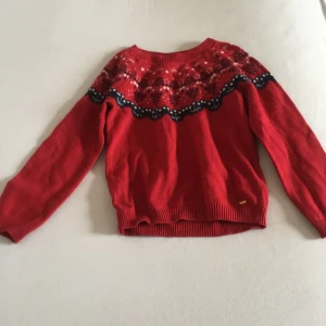 Röd stickad tröja med mönster - Säljer en röd stickad tröja med ett vackert mönster i svart och vitt runt halsen. Tröjan har långa ärmar och ribbstickade muddar vid ärmslut och nederkant. Perfekt för kyliga dagar och för julen!