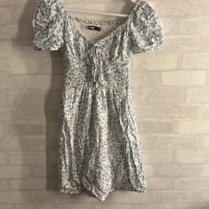 Blommig klänning från BikBok - Söt blommig klänning från BikBok. Den har korta puffärmar och en v-ringad design med knytning framtill. Klänningen är vit med blå blommor och har en smockad midja för en bekväm passform. Perfekt för sommaren!!