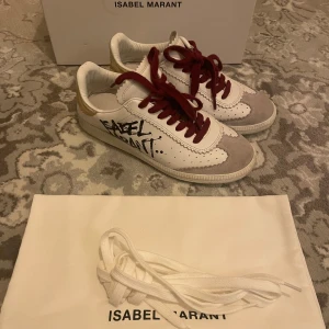 Vita sneakers från Isabel Marant - Snygga vita sneakers från Isabel Marant med röd snörning och svart text på sidan. Skorna har en stilren design med beige detaljer och kommer med extra vita skosnören. Perfekta för en trendig look! Storlek 36 ÄSLKAR dessa skor men säljer pga att de är lite små för mig 