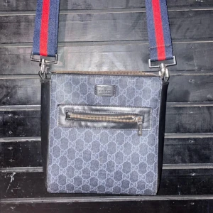 Axelväska från Gucci - Snygg axelväska från Gucci med ett klassiskt mönster i grått och svart. Väskan har en justerbar axelrem i blått och rött samt en praktisk dragkedja framtill. Perfekt för att bära med sig det viktigaste med stil.