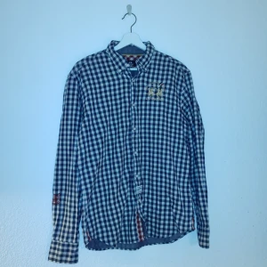 Rutig skjorta från La Martina - Säljer en snygg rutig skjorta från La Martina i blått och vitt. Skjortan har långa ärmar och ett broderat emblem på bröstet. Perfekt för en stilren look!