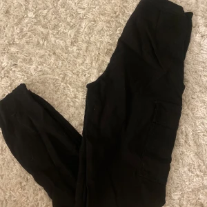 Svarta cargopants - Säljer ett par svarta cargopants med flera fickor och knappstängning. Perfekta för en avslappnad stil och praktiska med sina många fickor.
