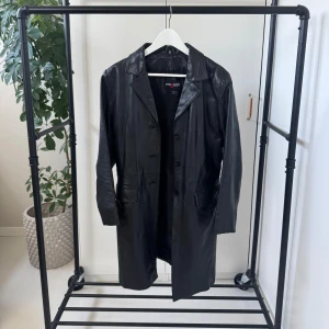 Svart skinnkappa - Snygg svart skinnkappa med klassisk krage och knappar framtill. Rocken har långa ärmar och två fickor framtill. Perfekt för en stilren look.