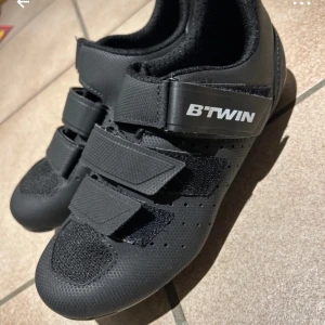 Svarta cykelskor från B'TWIN - Snygga svarta cykelskor från B'TWIN med tre justerbara kardborreband för optimal passform. Skorna har en stilren design och är perfekta för cykling. Tillverkade i syntetmaterial för hållbarhet och komfort.