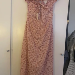 Blommig långklänning med knytning - Charmig långklänning i rosa med blommigt mönster. Klänningen har smala axelband och en fin knytning i ryggen. Perfekt för somriga dagar med sin luftiga design.