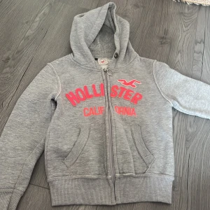Grå hoodie från Hollister - Säljer en grå hoodie från Hollister med rosa text och logga på framsidan. Den har en dragkedja och en mysig huva.🩶